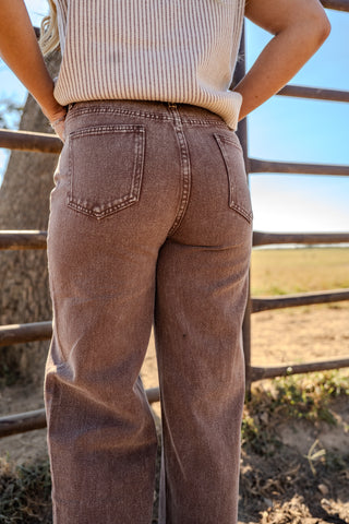 Person wearing brown jeans and a beige top standing in front of a wooden fence with a natural background.