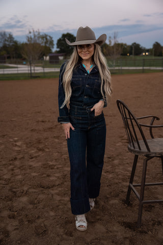 Woman in a cowboy hat and dark outfit standing in an open field with a chair nearby.