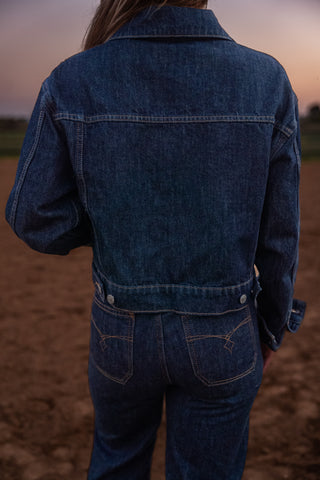 Person wearing a blue denim jacket and jeans standing in a field.