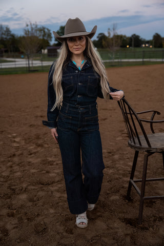 Woman in cowboy hat and dark outfit standing in a dirt field with a chair.