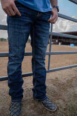 Person wearing blue jeans and a blue shirt standing near a metal fence with a dirt field and cloudy sky in the background.