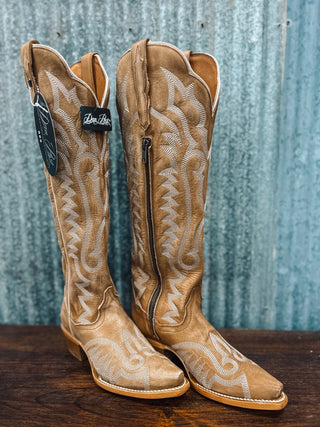 Pair of tan cowboy boots with intricate white embroidery on a wooden surface and blue textured background.