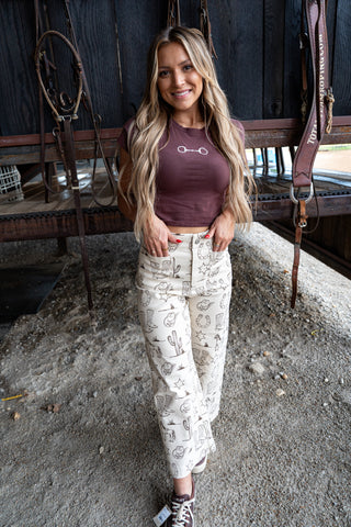 Woman wearing a maroon top and white patterned pants standing in an indoor setting with horse equipment.