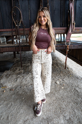 Woman standing in a barn wearing a brown top and patterned pants.