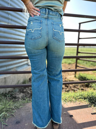 Person wearing blue jeans standing in a rural setting with a metal building and fence in the background.