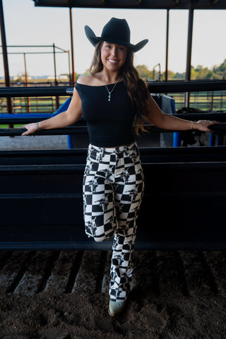 Woman wearing a black cowboy hat, black top, and checkered pants standing in an indoor arena.