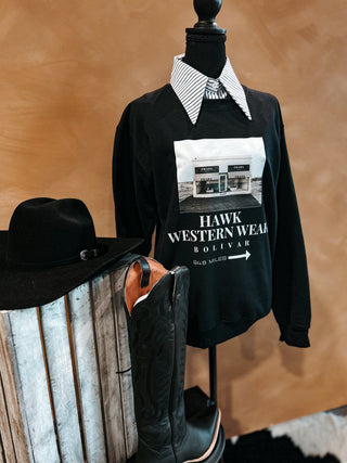 Black sweatshirt with graphic design on a mannequin, next to cowboy boots and a hat against a wooden background.