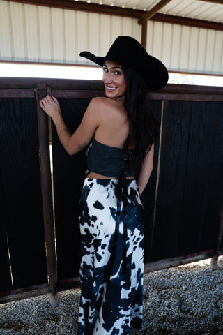 Woman wearing a black cowboy hat and cow print pants standing in a stable.