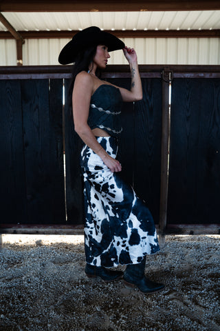 Woman in a strapless dress with cow print and black cowboy hat standing in a barn.