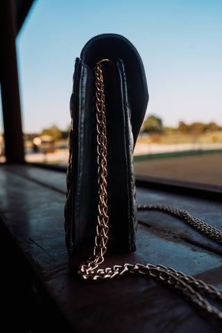 Black handbag with a gold chain strap on a wooden surface with a blurred outdoor background