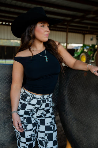 Woman wearing a black top and checkered pants standing next to a fence with a barn in the background.