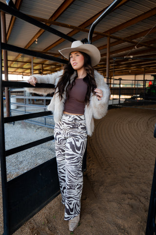 Woman in a barn wearing a white cowboy hat, brown top, and zebra print pants.
