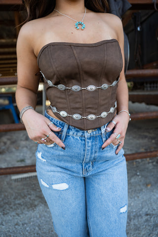 Person wearing a brown strapless top with silver belt and blue jeans outdoors.