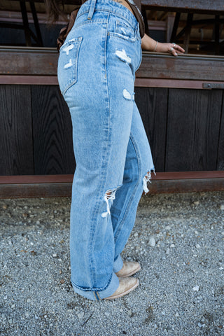 Person wearing light blue jeans with rips standing on a gravel surface.