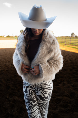 Person wearing a white fur coat, zebra print pants, and a white cowboy hat in an outdoor setting.