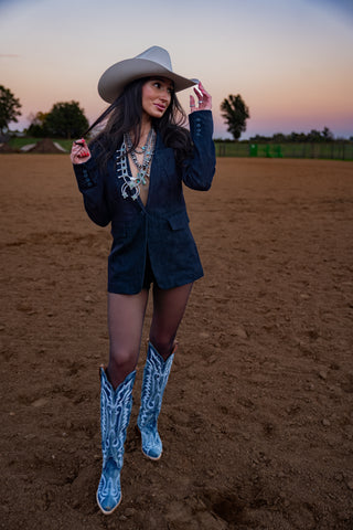 Woman in a blazer, cowboy hat, and blue cowboy boots standing on a dirt field.