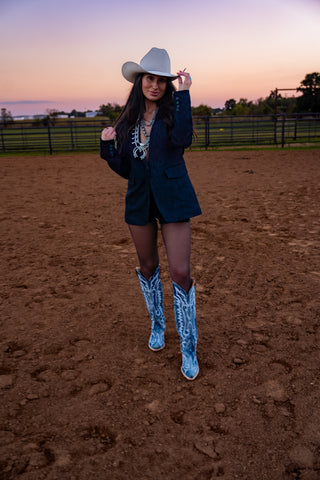 Woman in a cowboy hat and blue cowboy boots standing in a dirt field with a sunset sky.