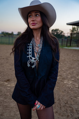 Woman wearing a cowboy hat and dark blazer with layered necklaces in an outdoor setting