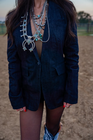 Person wearing a dark blue blazer with layered necklaces in an outdoor setting