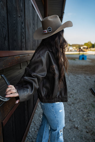Person wearing a cowboy hat and leather jacket leaning against a wooden structure.