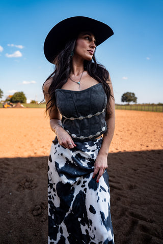Woman in a black cowboy hat and cow print skirt standing in an open field.