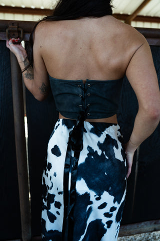 Person wearing a black corset top and cow print pants, standing against a dark background.