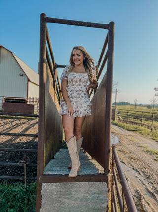 Woman in a floral dress and beige boots standing on a metal staircase with a barn and open field in the background.