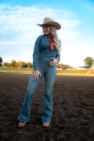 Woman in a denim outfit with a cowboy hat and scarf standing on a dirt field.