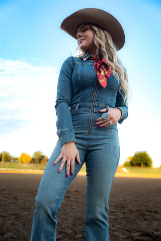 Woman in a denim outfit with a cowboy hat and red bandana standing in an open field.