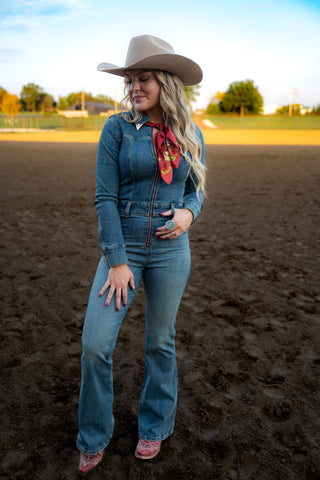 Woman in blue outfit with cowboy hat and red bandana standing on a dirt field.