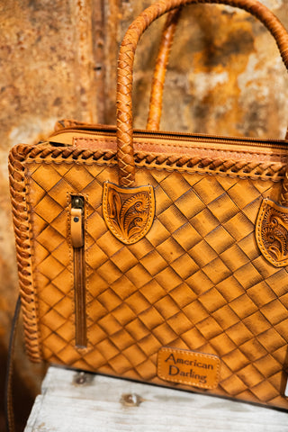 Brown leather handbag with braided handle and 'American Darling' branding on a rustic wooden surface.