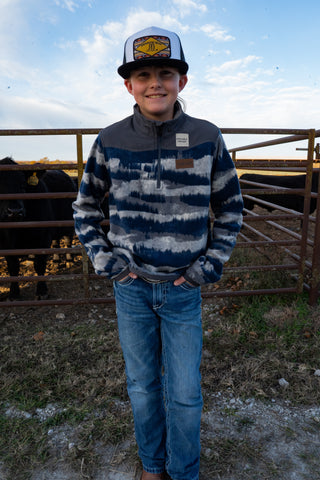 Young boy wearing a striped sweater and cap standing in front of a fence with livestock in the background.