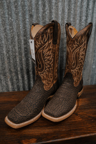 Pair of brown cowboy boots with intricate patterns on a wooden surface and corrugated metal background.