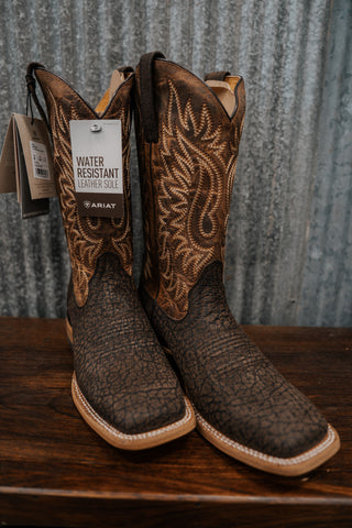 Pair of brown cowboy boots with intricate designs on a wooden surface and metal wall background.