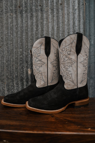 Pair of black cowboy boots with white tread pattern on a wooden surface and corrugated metal background.
