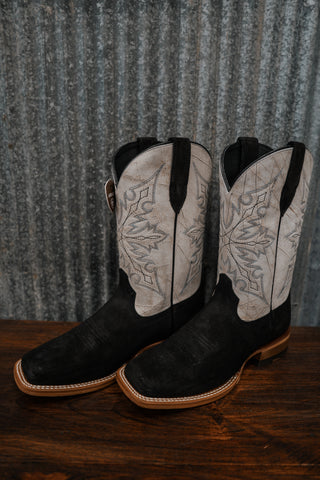 Pair of black cowboy boots with white embroidery on a wooden surface and corrugated metal background.
