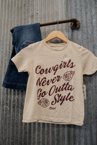 Beige t-shirt with 'Cowgirls Never Go Outta Style' text on a hanger against a rustic metal wall.