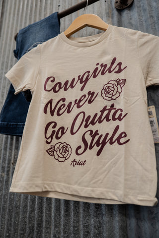 Beige t-shirt with 'Cowgirls Never Go Outta Style' text on a hanger against a textured background.