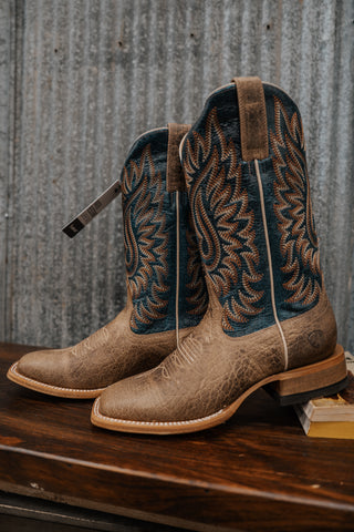 Pair of brown cowboy boots with intricate designs on a wooden surface and metal wall background