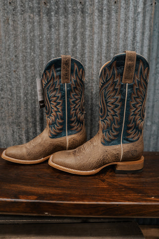 Pair of tan cowboy boots with intricate designs on a wooden surface and metal wall background