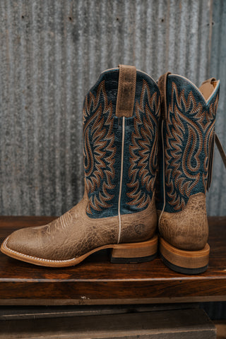 Pair of brown cowboy boots with intricate designs on a wooden surface and metal wall background