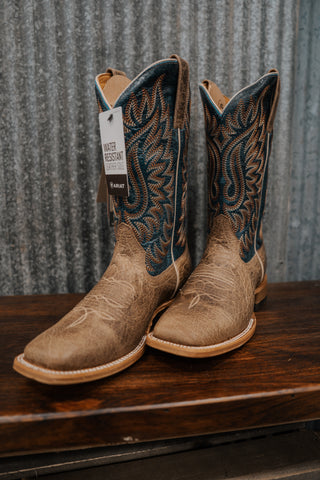 Pair of tan cowboy boots with intricate designs on a wooden surface and metal background