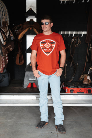 Man wearing an Ariat red t-shirt standing in a garage with tools and equipment.