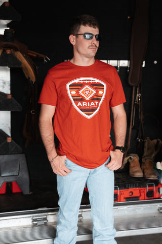 Man wearing a red Ariat t-shirt in an indoor setting with equipment and boots in the background