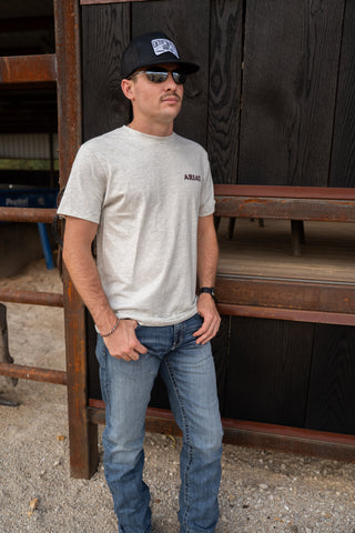 Man wearing a cap, sunglasses, and a t-shirt standing in front of a wooden structure.