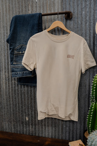 Beige t-shirt with a logo on a hanger against a corrugated metal wall.