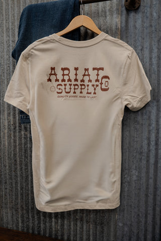 Beige t-shirt with 'Ariat Supply' logo on a hanger against a corrugated metal background