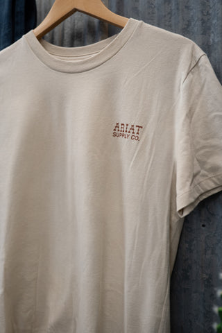 Beige t-shirt with Ariat Supply Co. logo on a hanger against a textured wall.