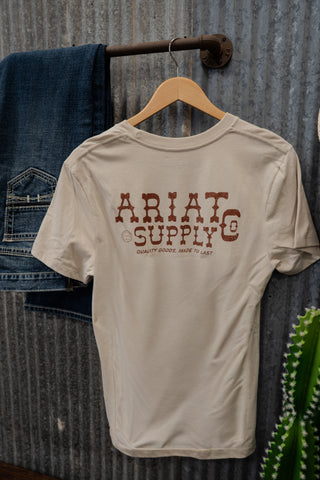 Beige t-shirt with 'Ariat Supply Co' text on a hanger against a rustic background.