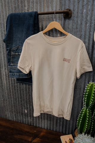 Beige t-shirt with a logo on a wooden hanger against a corrugated metal wall.